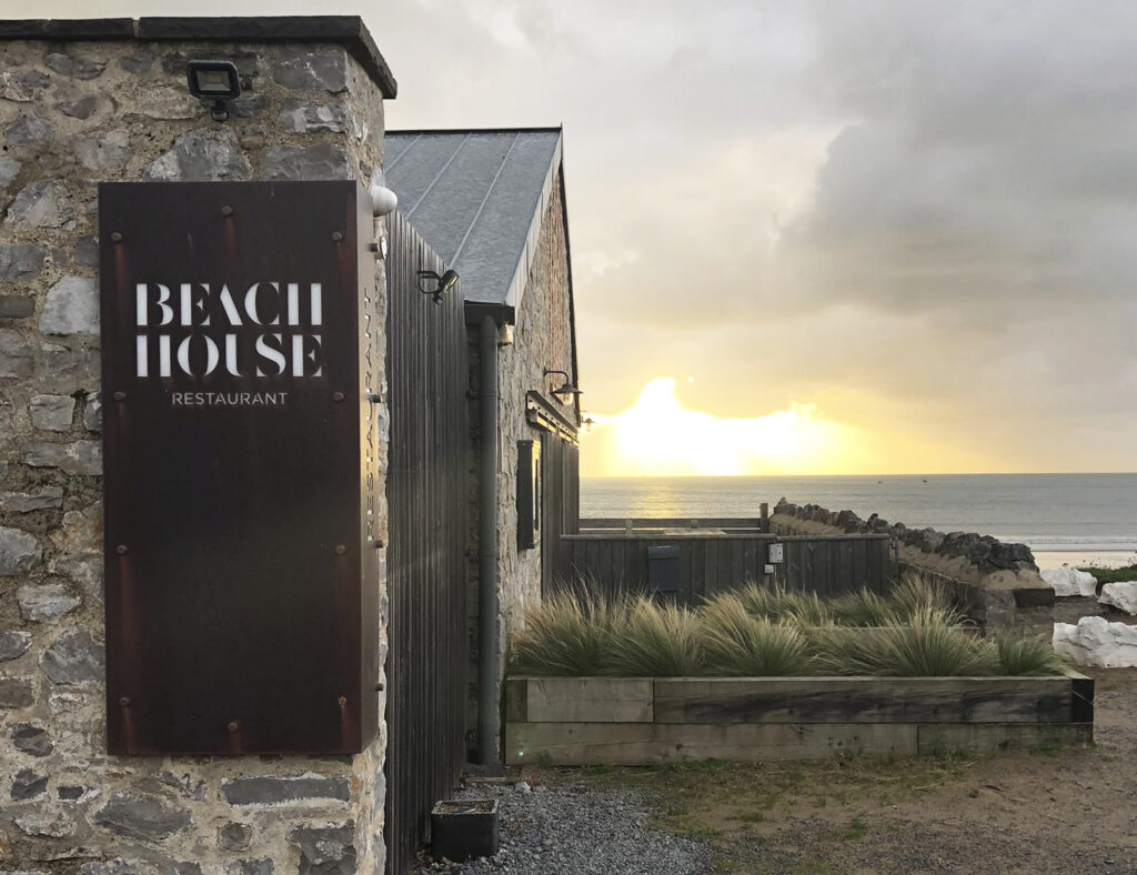 A sign with the Beach House brand mark is attached to the wall of this high-end restaurant, with the sun setting over the sea in the background