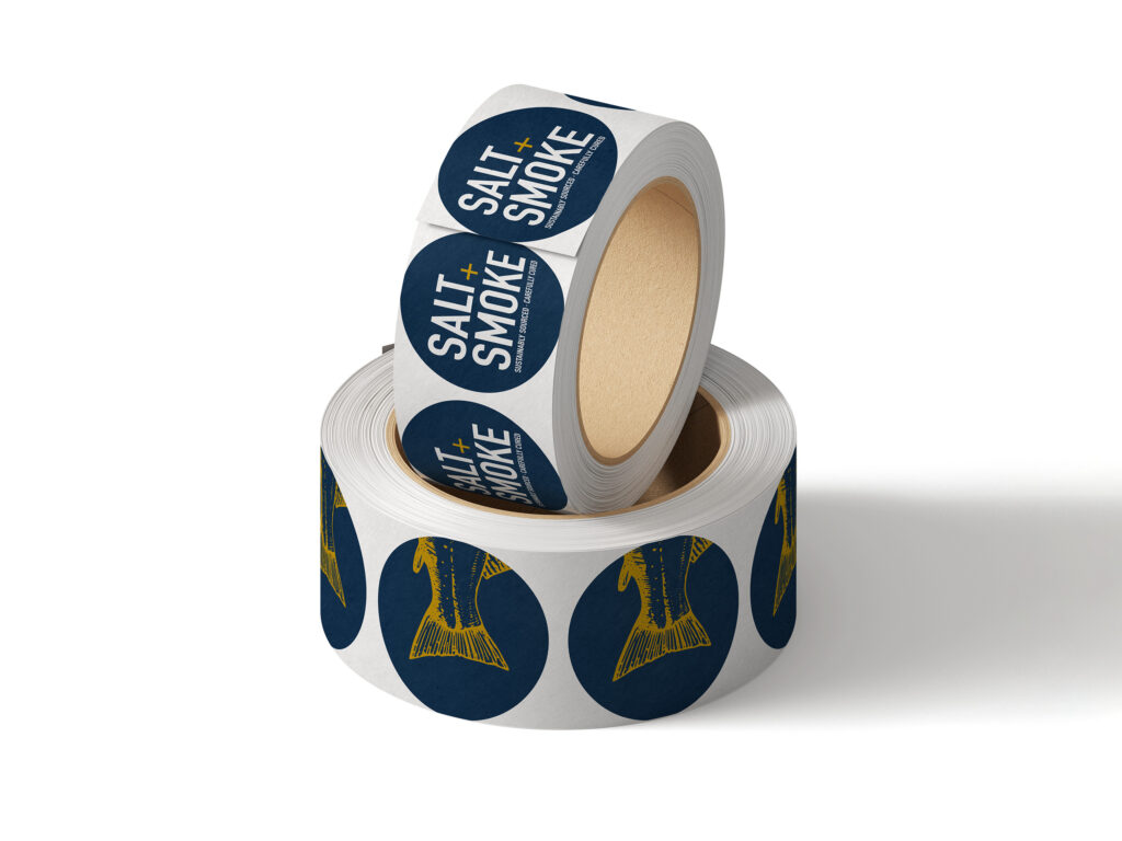 Sticker design for packaging, two rolls of round stickers with different designs; one is navy blue with the white Salt + Smoke logo, the other is navy blue with a gold salmon tail illustration