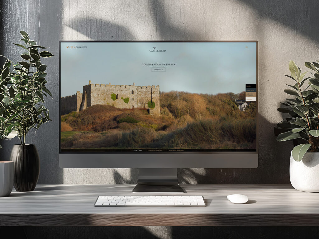 A desk with an imac showing the homepage of the Castlemead hotel website making use of their branding and strong imagery
