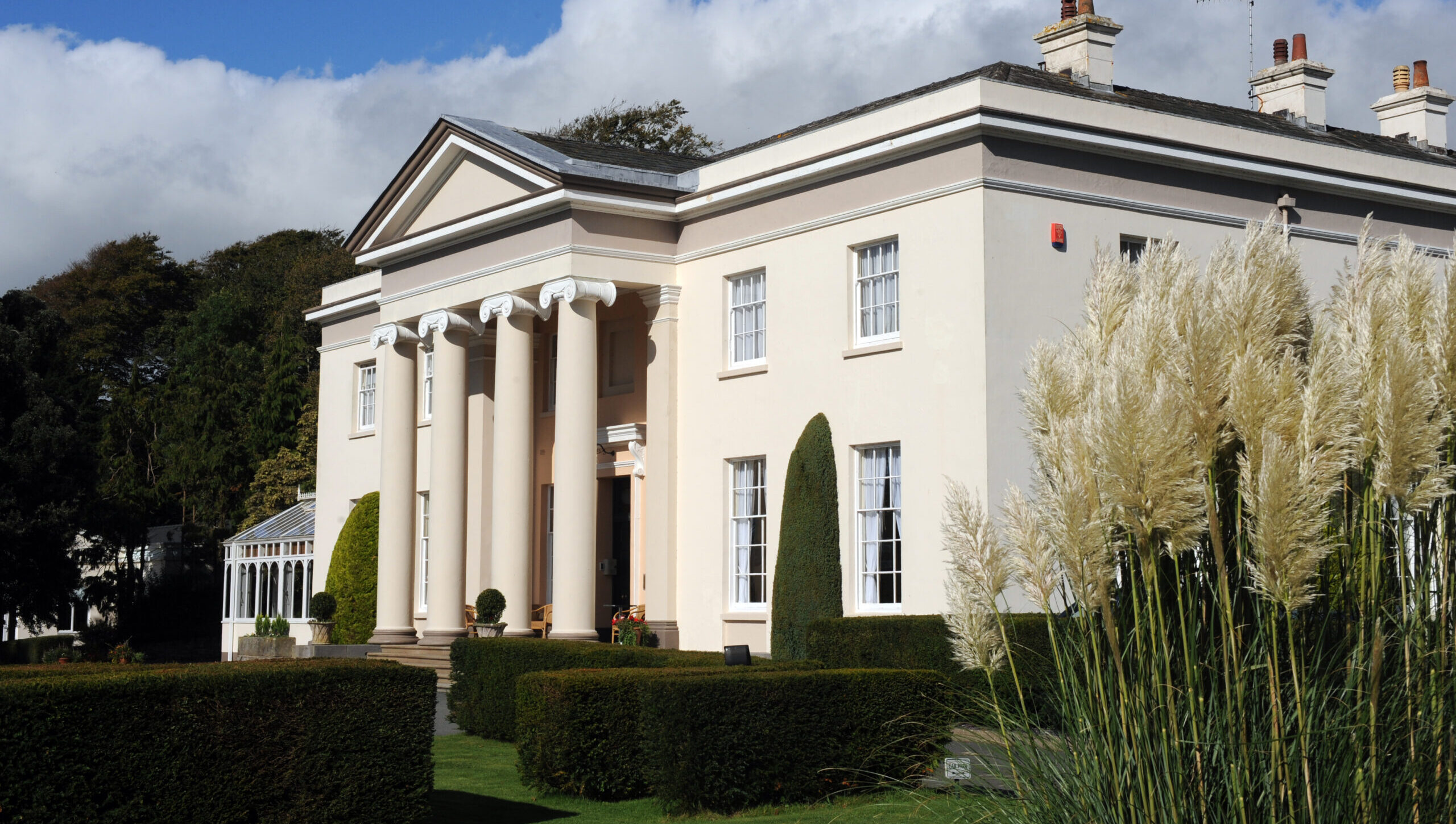 An exterior shot of a Georgian Mansion painted cream with greenery in the foreground and a cloudy blue sky