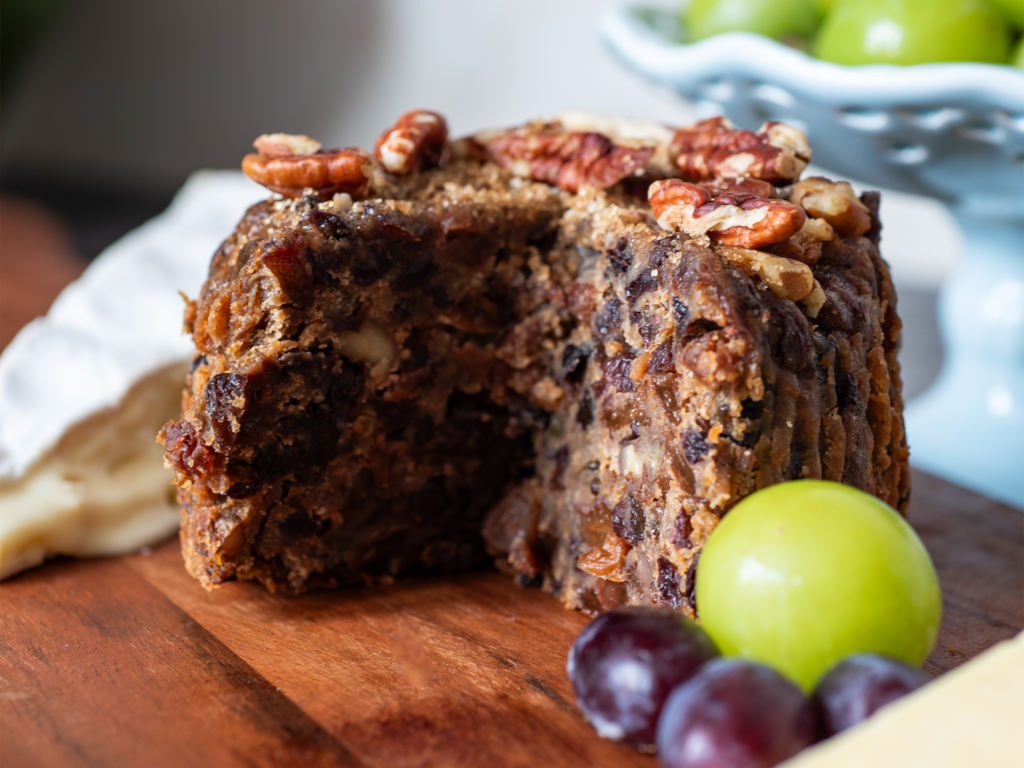 A detailled image of a round cake called Torsy Morsy Cake which has a slice cut out of it and some grapes in the forefront of the chopping board