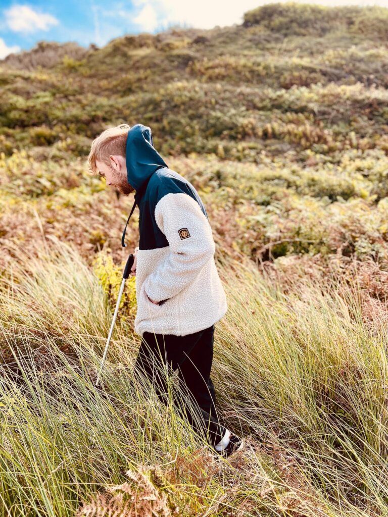 A man wearing a white and teal hooded jacket walking among long grass in sand dunes holding a litter picker
