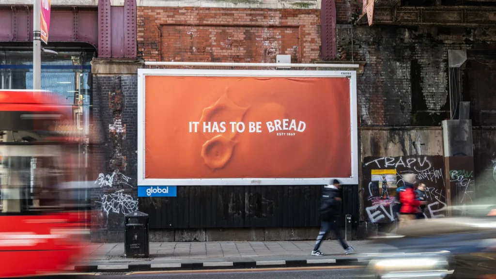 A billboard on a brick wall with a red London bus in the left side of the image, on the billboard is a picture of Heinz sauce with the words 'It has to be Bread' written on it